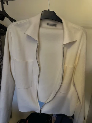 Krämvit ziptröja från Åhléns - Stilren krämvit ziptröja från Åhléns med två stora bröstfickor och klassisk krage. Tröjan har lång ärm och är tillverkad i ett mjukt, stickat material som känns skönt mot huden. Perfekt att slänga på sig över en t-shirt för en avslappnad look.