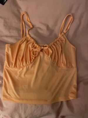 Ljus orange linnetopp med rosett - Supersöt ljus orange linnetopp med smala axelband och en liten rosett framtill. Toppen har en lätt rynkad detalj vid bysten och är croppad i modellen. Perfekt för varma dagar och enkel att styla med jeans eller kjol.