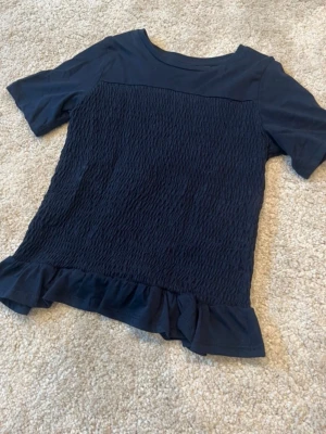 Mörkblå topp med volang från Gina Tricot - Supersöt mörkblå topp från Gina Tricot med korta ärmar, smockad framsida och volangkant nedtill. Perfekt för dig som gillar en enkel men ändå trendig stil. Tillverkad i mjukt material som känns skönt mot huden.