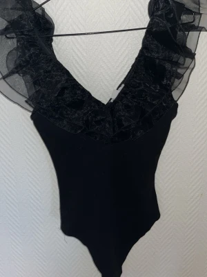Svart body med volangdetaljer - Snygg svart body med djup V-ringning och stora volanger i transparent mesh längs axlar och urringning. Jätte bra skick och storlek s men passsar xs också.