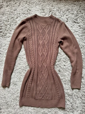 Brun kabelstickad lång tröja - Säljer en brun kabelstickad lång tröja med snyggt mönster framtill. Tröjan har rund halsringning, långa ärmar och ribbade muddar. Perfekt för lager på lager och riktigt mysig till hösten.