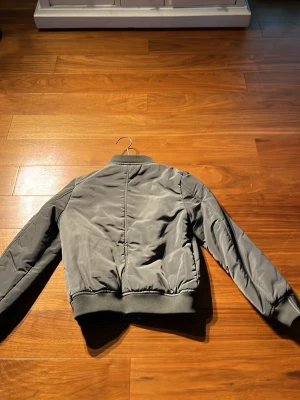 Grå bomberjacka med fickor - Snygg grå bomberjacka med dragkedja framtill och två fickor med tryckknappar. Jackan har ribbade muddar vid ärmslut och midja samt en extra ficka med dragkedja på ena ärmen. Klassisk bombermodell i vadderat material.