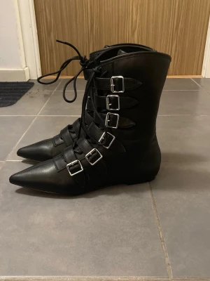 Svarta winklepickers strl 39 - Säljer ett par helt nya svarta Winklepickers/pikes från Strange Cvlt i strl 9 (USA, man) vilket jag upplevde som en strl 39 (EU, kvinna). Spännena är inte riktiga utan de spänns fast med kardborrband. Snörena går att spänna åt. Skorna är helt oanvända då jag endast provat de en gång inomhus. Skorna är perfekta för dig som är goth och vill äga ett par klassiska gothskor. Frakt ingår ej