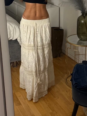 Vit långkjol med broderade detaljer - Säljer en superfin vit långkjol med broderade hålmönster och volangdetaljer. Kjolen har elastisk midja och är gjord i ett lätt bomullstyg som passar perfekt till varmare dagar. Perfekt att styla med en crop top eller en enkel t-shirt.