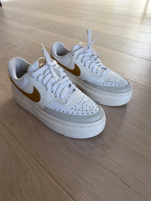 Nike Air Force 1 Sage Low vit/guld - Säljer ett par vita Nike sneakers med guldfärgad swoosh och grå detaljer. Tillverkade i läder och textil, perfekta för en clean och sportig look. Skorna är väldigt bekväma men använder tyvärr inte dem längre. Dessa skor finns inte längre att få tag på✨🔥