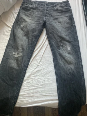 Svarta slitna jeans med raka ben - Säljer ett par svarta jeans med slitningar på båda benen och klassisk femficksmodell. Jeansen har raka ben och en cool tvättad look. Materialet är denim och bakfickan har en diskret söm. Perfekta för en avslappnad streetstil.