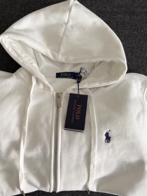 Vit hoodie från Polo Ralph Lauren - Snygg vit hoodie från Polo Ralph Lauren med dragkedja framtill och klassisk blå logga broderad på bröstet. Hoodien har huva med dragsko, ribbade muddar och fickor på sidorna. Perfekt för en clean och stilren look.