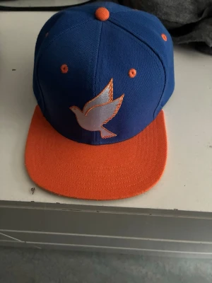 Blå och orange keps från Galagowear - Säljer en snygg snapback-keps från Galagowear i blått med orange skärm och detaljer. Framtill finns en broderad vit duva med orange kant. Justerbar baktill med broderad logga. Perfekt för dig som vill sticka ut med färg och stil. Kan diskutera pris 