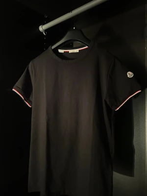 Svart Moncler t-shirt med logga - Snygg svart t-shirt från Moncler med klassisk rund hals och korta ärmar. Ärmsluten har rödvita detaljer och Moncler-loggan sitter på vänster ärm. Tillverkad i mjuk bomull för skön känsla och stilren look. 