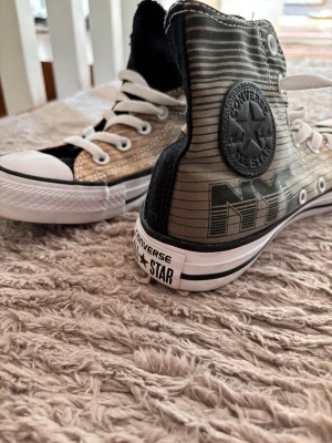 Converse NYC High Top sneakers - Snygga Converse All Star high tops med NYC-tryck på sidan. Skorna har svart och beige randigt tyg, vita gummisulor och tåhättor samt vita skosnören. Klassisk Converse-logga på ankeln och cool streetstyle-vibe. Perfekta för dig som vill sticka ut. Storleken: 37,5