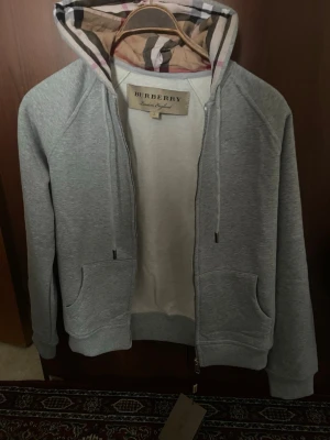 Grå zip hoodie från Burberry - Snygg grå zip hoodie från Burberry med klassiskt rutigt foder i huvan och dragsko. Hoodien har två fickor framtill och ribbade muddar vid ärmar och nederkant. Perfekt för en chill och stilren look. XL men passar s.