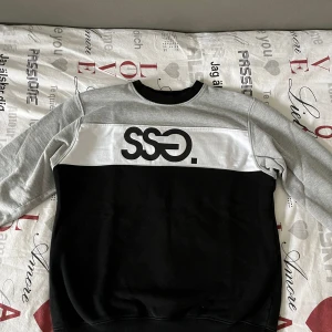 Svart och grå sweatshirt från SSG - Säljer en snygg svart och grå sweatshirt från SSG med deras logga tryckt på framsidan. Tröjan har långa ärmar och en bekväm passform. Perfekt för en avslappnad stil.