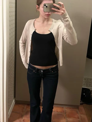 Beige tunn cardigan med knappar - Snygg och enkel beige cardigan med rund halsringning och knäppning framtill. Modellen är tunnstickad och har långa ärmar samt ribbade muddar vid ärmslut och nederkant. Perfekt att slänga över ett linne eller t-shirt.