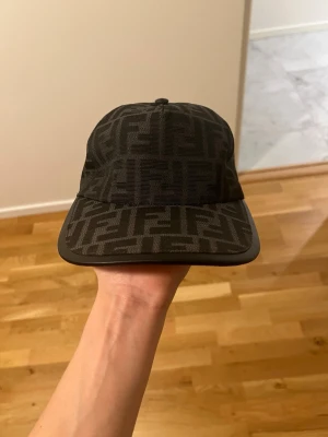 Svart keps från Fendi äkta  - Snygg svart keps från Fendi med det klassiska FF-logomönstret över hela kepsen. Kepsen har böjd skärm och justerbar rem baktill. Tillverkad i textilmaterial som ger en exklusiv känsla.