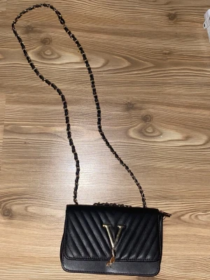 Svart dam väska - Snygg svart axelväska med quiltat mönster och guldfärgad kedjerem. Väskan har ett stort V i metall framtill och en elegant tofsdetalj. Perfekt för dig som vill ha en trendig och stilren accessoar. 