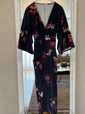 Mörkblommig omlottklänning i sammet - Långklänning morgonrock i mjuk sammet med mörk botten och blommigt mönster i lila, rosa och vinröda toner. Omlottmodell med djup v-ringning och vida ärmar. Perfekt för dig som vill ha en statementklänning med lyxig känsla.