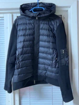 Svart pufferjacka från Moncler - Säljer en svart pufferjacka från Moncler med huva och dragkedja framtill. Jackan har stickade ärmar, två fickor med dragkedja och en extra ficka på ena ärmen. Klassisk Moncler-logga på armen och quiltad design på kroppen. Perfekt för kalla vinterdagar.