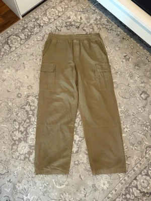Beiga cargobyxor med fickor - Säljer ett par beiga cargobyxor med elastisk midja och stora sidofickor. Byxorna har raka ben och är tillverkade i ett mjukt bomullsliknande material. Perfekta för en avslappnad och trendig look.