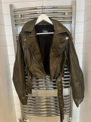 Olivgrön skinnjacka från Zara - Snygg olivgrön skinnjacka (konstläder) från Zara med breda slag, asymmetrisk dragkedja och bälte i midjan. Jackan har coola detaljer som silverfärgade knappar och dragkedjor samt markerade sömmar. 
