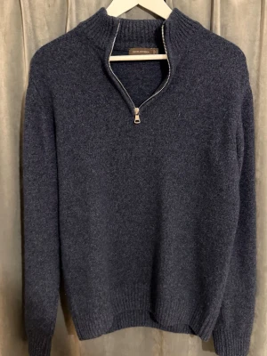 Mörkblå stickad tröja med halvzip - Mörkblå stickad tröja från Oscar Jacobson med halvzip och  krage. Tröjan har ribbstickade muddar vid ärmslut och nederkant samt en mjuk, lite fluffig yta. Perfekt för lager på lager och enkel att matcha med olika outfits.