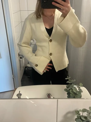 Krämvit kofta med knappar - Säljer en krämvit kofta med stora bruna knappar och v-ringad hals. Koftan har långa, vida ärmar och en rak passform. Perfekt att slänga över en topp för en clean och enkel look.