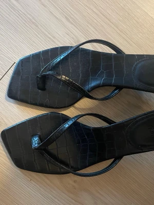 Svarta sandaler med krokopräglad yta H&M - Säljer ett par svarta sandaler från H&M med trendig fyrkantig tå och krokopräglad yta. Remmarna är tunna och minimalistiska, vilket ger en clean look. Perfekta för dig som gillar stilrena detaljer och vill ha något som sticker ut.