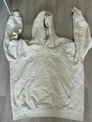 Vit hoodie från Sail Racing - Säljer en stilren vit hoodie från Sail Racing med broderad text och siffran 77 på bröstet. Hoodien har en stor känguruficka framtill, dragsko i huvan och ribbade muddar. Perfekt för dig som gillar sportig och clean stil.