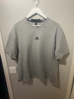 Grå Adidas t-shirt i bomull med prislappen kvar - Snygg grå t-shirt från Adidas med klassisk logga på bröstet. T-shirten har rund halsringning och korta ärmar. Tillverkad i mjuk bomull för en skön och avslappnad look. Passar till all form av träning.