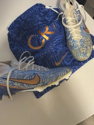 Nike CR7 blåa fotbollsskor - Snygga fotbollsskor från Nike CR7-kollektionen i blått och vitt med guldiga detaljer och mönster. Skorna har vita skosnören, guldigt Swoosh och CR7-logga på sidan. Yttermaterialet är syntet och sulan är anpassad för spel på gräs.