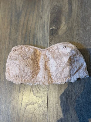 Ljusrosa bandeau spets-bh - Säljer en ljusrosa bandeau-bh i mjuk spets med blommönster. Bh:n är axelbandslös och har elastisk kant nedtill för extra stöd. Insidan är fodrad i ett mjukt tyg för bekväm känsla. Perfekt under toppar med bara axlar.