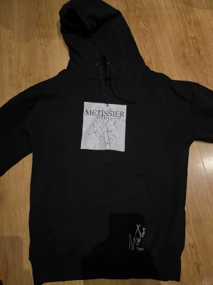 Svart hoodie från Métissier Paris - Svart hoodie från Métissier Paris med stor huva och snörning. På bröstet finns en vit patch med sprucket tryck och logga. Klassisk känguruficka framtill och diskret signaturdetalj nedtill. Perfekt för en avslappnad streetwear-look.