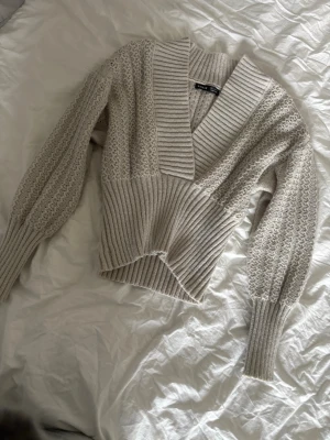 Beige stickad tröja från Gina Tricot - Säljer en beige stickad tröja från Gina Tricot med bred ribbad mudd i midjan och vid ärmsluten. Tröjan har djup v-ringning, ballongärmar och ett snyggt strukturmönster. Perfekt för dig som gillar en trendig och mysig look.