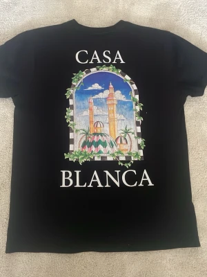 Svart Casablanca t-shirt - Snygg svart t-shirt från Casablanca med färgglatt tryck av arkitektur och palmer på ryggen samt mindre tryck framtill. T-shirten har korta ärmar och rund hals. Perfekt för dig som vill sticka ut med en unik design. 