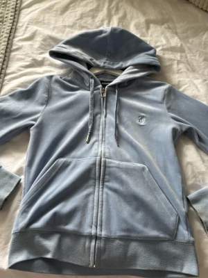 Ljusblå velourhoodie Juicy Couture - Säljer en ljusblå velourhoodie från Juicy Couture med dragkedja, huva och fickor framtill. Klassisk logga broderad på bröstet och mjukt material som känns lyxigt. Perfekt för en chill och trendig look.