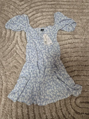 Blommig ljusblå klänning från Gina Tricot - Supergullig ljusblå kortklänning från Gina Tricot med vitt blommönster. Klänningen har puffiga kortärmar, slimmad midja och lätt utställd kjol. Perfekt nu tiöl sommarens alla tillfällen. Kan justeras och knytas vid byst. Tillverkad i mjuk bomull för en skön känsla. Aldrig använd, prislapp på. Säljs inte längre. Stlk xs. Skriv om funderingar❤️