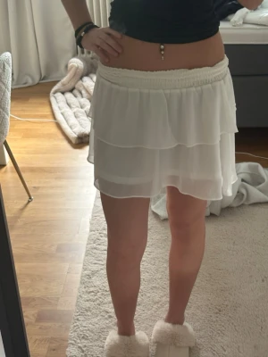 Vit volangkjol med resårmidja - Söt vit kortkjol med volanger och bred resår i midjan. Kjolen är gjord i ett lätt, flödande material som ger en luftig känsla. Perfekt för varma dagar och enkel att matcha med olika toppar.