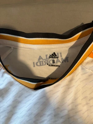 Real Madrid matchtröja Adidas S - Säljer en vit Real Madrid matchtröja från Adidas med svarta och gula detaljer. Tröjan har klubbmärke, Adidas-logga och sponsortryck på bröstet samt svarta ränder på sidorna. Tillverkad i lätt och ventilerande material med Climalite-teknologi.