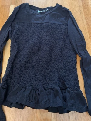 Svart långärmad topp med volang - Säljer min svarta tröja i storlek 156/164 från Gina då den knappt har kommit till användning. Svart långärmad topp med smockad kropp och volangkant nedtill. Toppen har rund halsringning och är tillverkad i ett mjukt material som känns bekvämt mot huden. Perfekt för dig som gillar en enkel men ändå detaljrik stil.