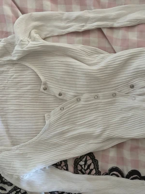 Vit ribbad långärmad topp - Säljer en vit ribbad långärmad topp med knappar framtill. Toppen har en djup u-ringning och är tillverkad i ett mjukt, stretchigt material som sitter snyggt på kroppen. Perfekt att styla med jeans eller kjol.