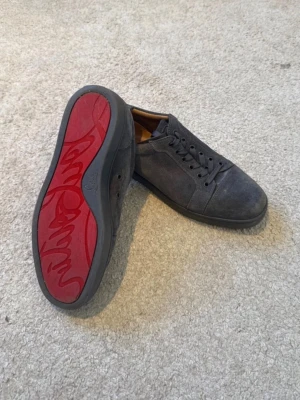 Gråa Louboutin sneakers i mocka - Säljer ett par gråa sneakers från Christian Louboutin i mjuk mocka med klassisk röd sula och diskret logga undertill. Skorna har lågt skaft, matchsnde snören och en stilren design som passar till det mesta. Box inkluderande extra snören mm medföljer. 