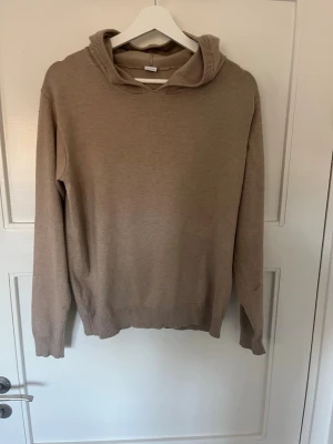 Beige stickad hoodie - Säljer en beige stickad hoodie med långa ärmar och ribbade muddar vid ärmslut och nederkant. Tröjan har en avslappnad passform och en mjuk huva. Perfekt för dig som gillar enkel och stilren design.