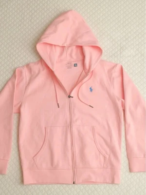  Ralph Lauren zip-up - Snygg ljusrosa hoodie med dragkedja från Polo Ralph Lauren. Klassisk design med huva och dragsnören, två fickor framtill och den ikoniska blå loggan broderad på bröstet. Tillverkad i mjuk bomull som känns skön mot huden. Perfekt för en chill och avslappnad stil.