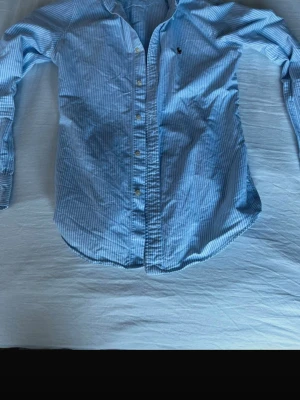 Blårandig skjorta från Ralph Lauren - Klassisk blårandig skjorta från Ralph Lauren med lång ärm och knappar framtill. Skjortan har en liten broderad logga på bröstet och är tillverkad i mjuk bomull. Perfekt för en stilren och avslappnad look.