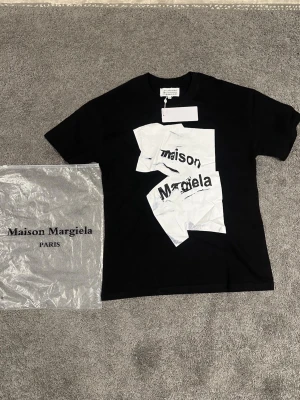 Maison Margiela svart t-shirt - Maison Margiela svart t-shirt med tryck framtill i vitt och grått. Klassisk passform med korta ärmar och rund hals. Tillverkad i mjuk bomull och levereras med originalpåse. Perfekt för dig som gillar streetwear och designerplagg.
