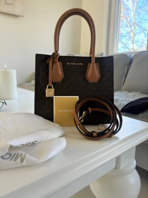 Michael Kors brun handväska - Snygg handväska från Michael Kors i mörkbrunt med logomönster och ljusbruna detaljer. Väskan har två handtag, avtagbar axelrem och guldfärgade metalldetaljer. Använd 1/2 gånger. 