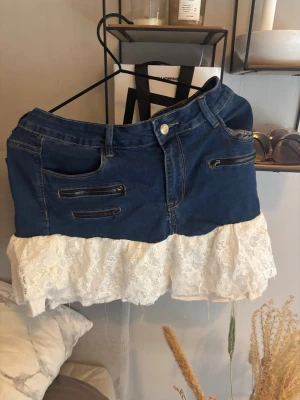 Jeanskjol med spetsvolang - Säljer en blå jeanskjol med flera dragkedjedetaljer framtill och en vit spetsvolang nedtill. Kjolen har klassiska jeansfickor och knappstängning i midjan. Perfekt för dig som gillar en mix av denim och romantiska detaljer.