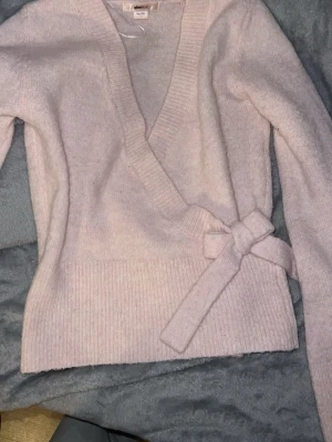 Ljusrosa omlottstickad tröja - Supersöt ljusrosa stickad omlottopp från Gina med knytband i midjan och djup v-ringning. 