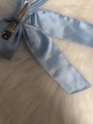 Ljusblå sidenrosett hårklämma - Supersöt hårklämma i ljusblått sidenliknande tyg, formad som en stor rosett med långa band. Metallklämma på baksidan gör den enkel att fästa i håret. Perfekt accessoar för att piffa till frisyren med en elegant touch.