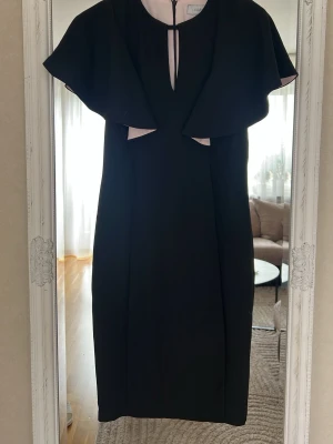 Svart klänning med cape från Coast - Stilren svart klänning från Coast med cape-liknande överdel och ljusrosa detaljer vid hals och midja. Klänningen har en elegant, rak passform och nyckelhålsöppning fram. Perfekt för dig som vill ha en unik och chic look.