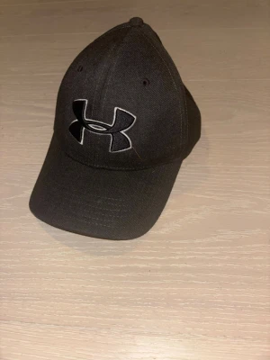 Svart keps från Under Armour - Snygg svart keps från Under Armour med böjd skärm och stort broderat logotyp framtill. Klassisk modell med ventilerande hål och justerbar passform. Perfekt för dig som vill ha en stilren och sportig look.
